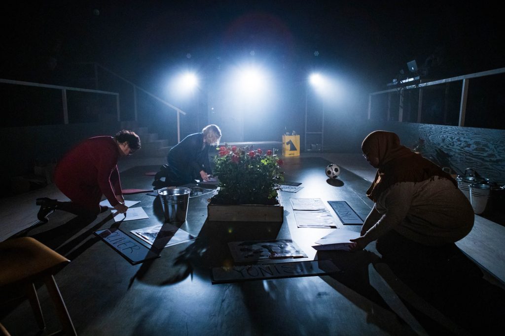 Maria, Ellie och Iman sitter på knä i scenografin och skriver på skyltar. I scenografin syns också en planterad rosbuske, en gul kasse och en fotboll.